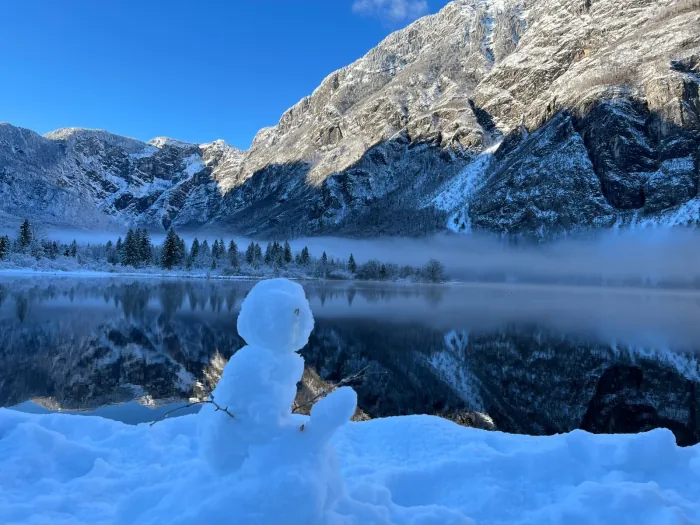 A snowman by the shore of Lake Bohinj, mountains beyond